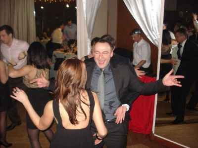 Rozbawiony mężczyzna w garniturze i krawacie, z szeroko rozłożonymi ramionami, tańczy na parkiecie podczas imprezy. W tle widoczni inni bawiący się goście i elementy dekoracji sali.