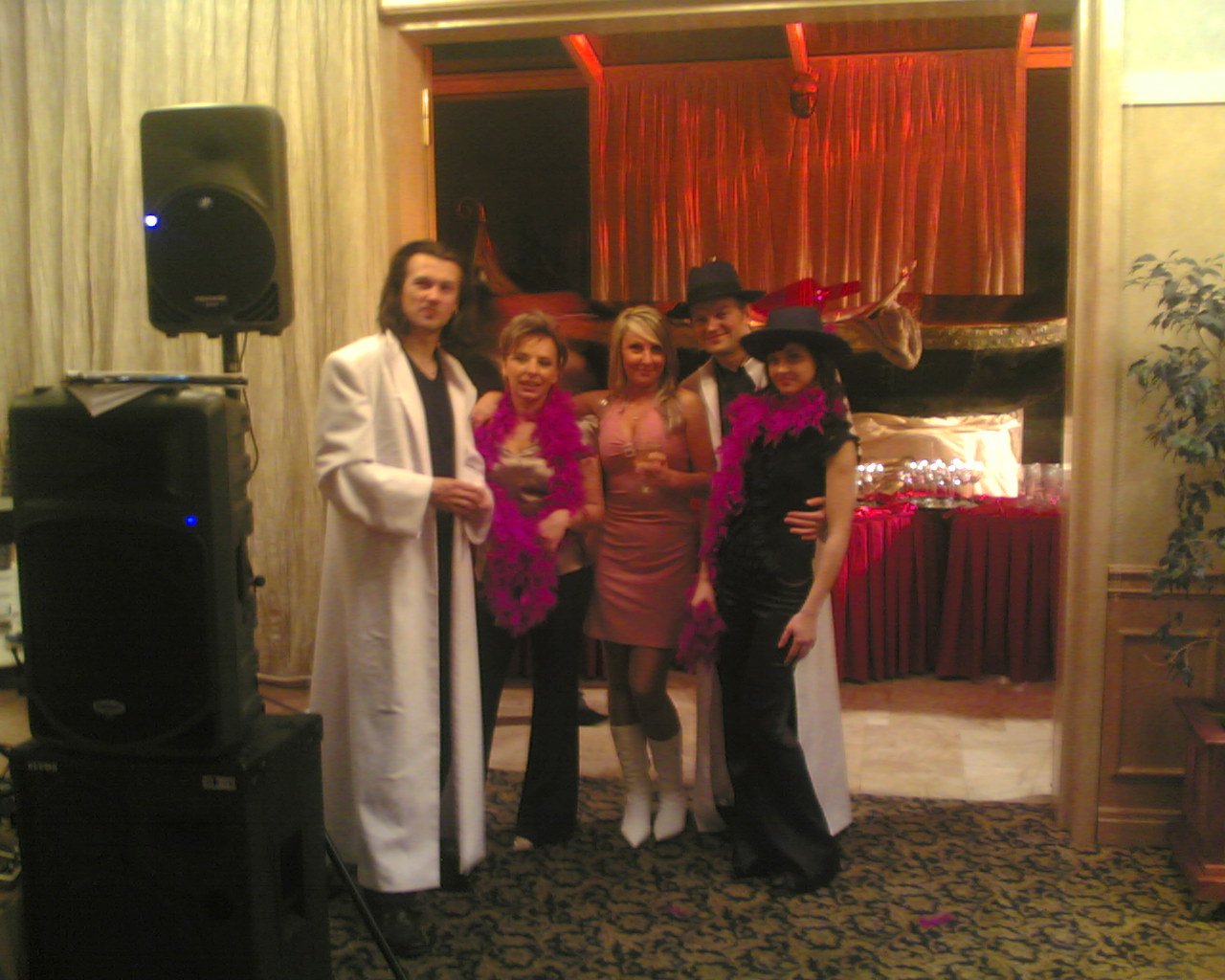 Grupa osób w strojach tematycznych z boa, ustawiona obok nagłośnienia i oświetlenia na tle dekoracji scenicznej z czerwonymi zasłonami i bufetem.