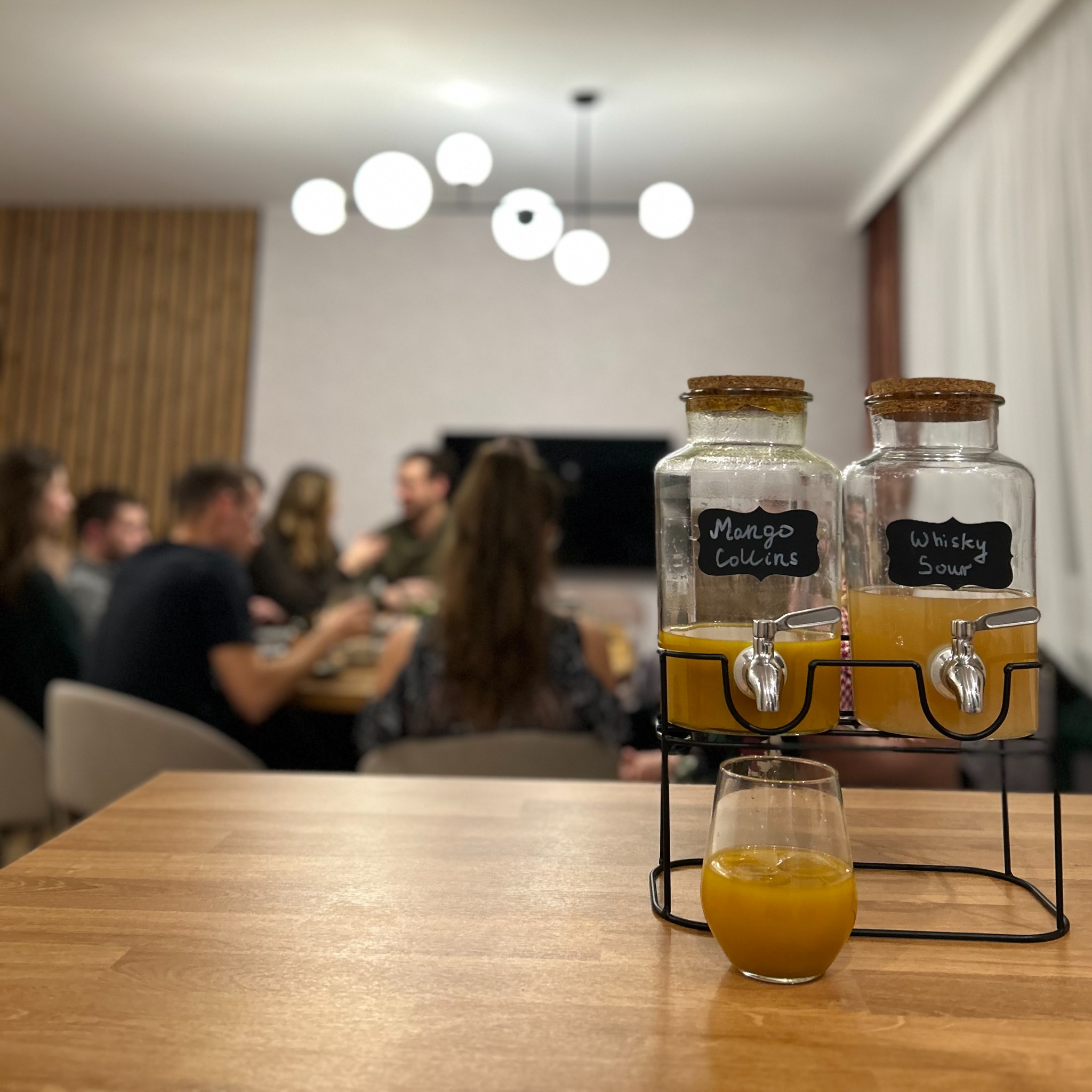 Dwa szklane dystrybutory napojów z napisami Mango Collins i Whisky Sour, stojące na drewnianym stole, w tle grupa osób siedząca przy stole w nowoczesnym wnętrzu.
