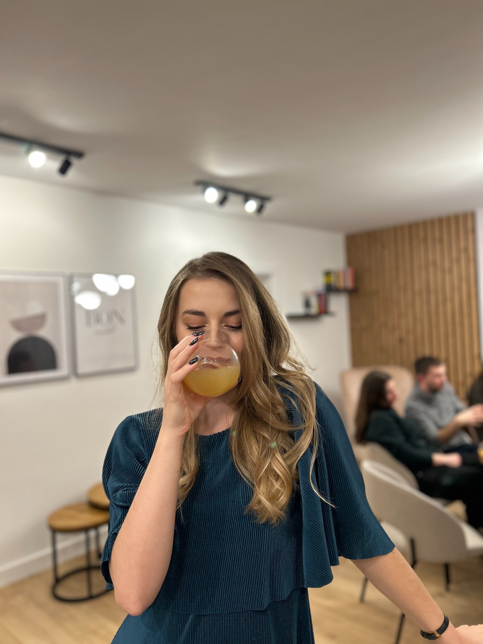 Uśmiechnięta kobieta w turkusowej sukience pije żółty napój z okrągłej szklanki podczas spotkania towarzyskiego w jasnym wnętrzu.