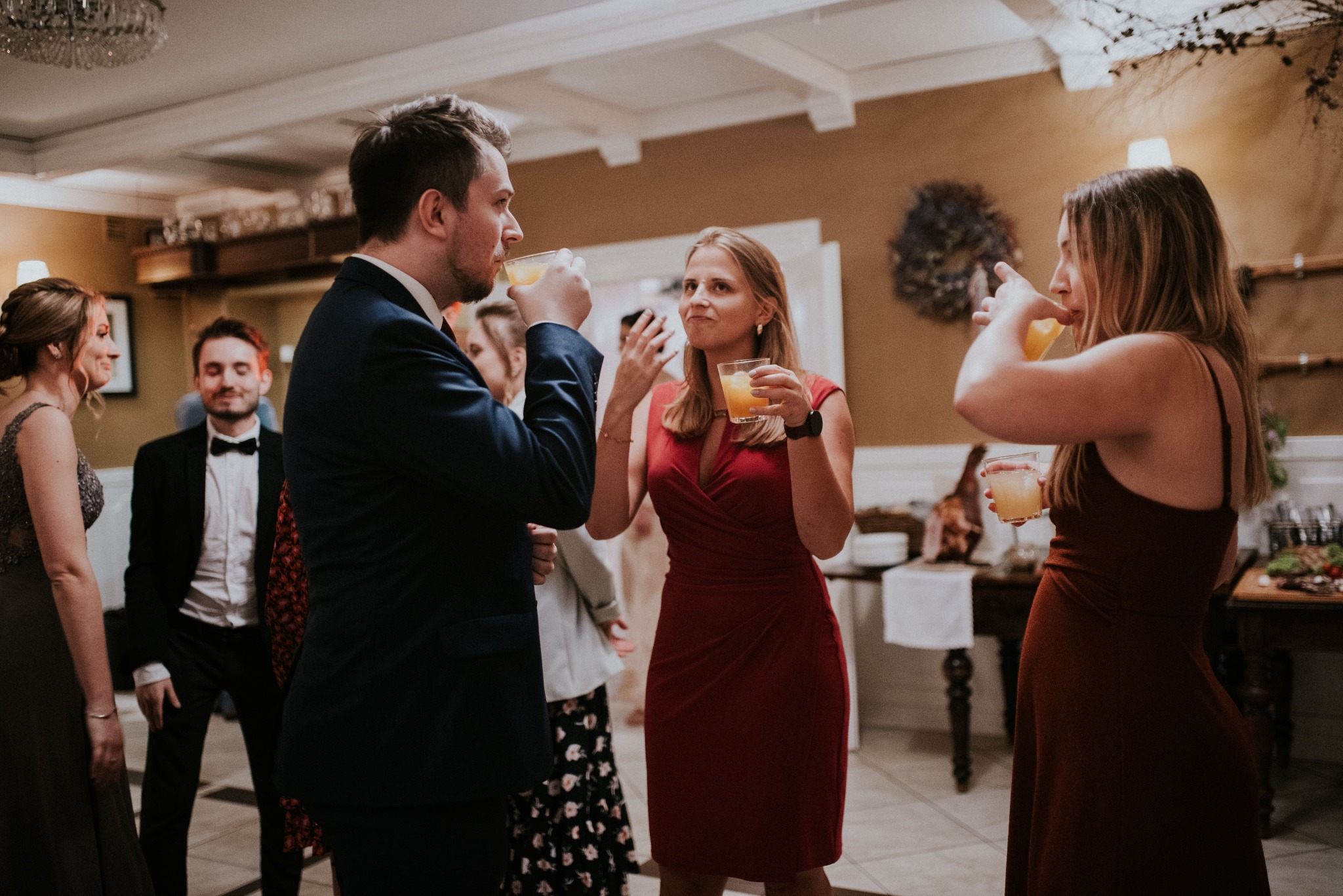 Grupa osób w eleganckich strojach wznosi toast pomarańczowymi drinkami podczas przyjęcia w jasnym wnętrzu z żyrandolem i dekoracjami kwiatowymi.