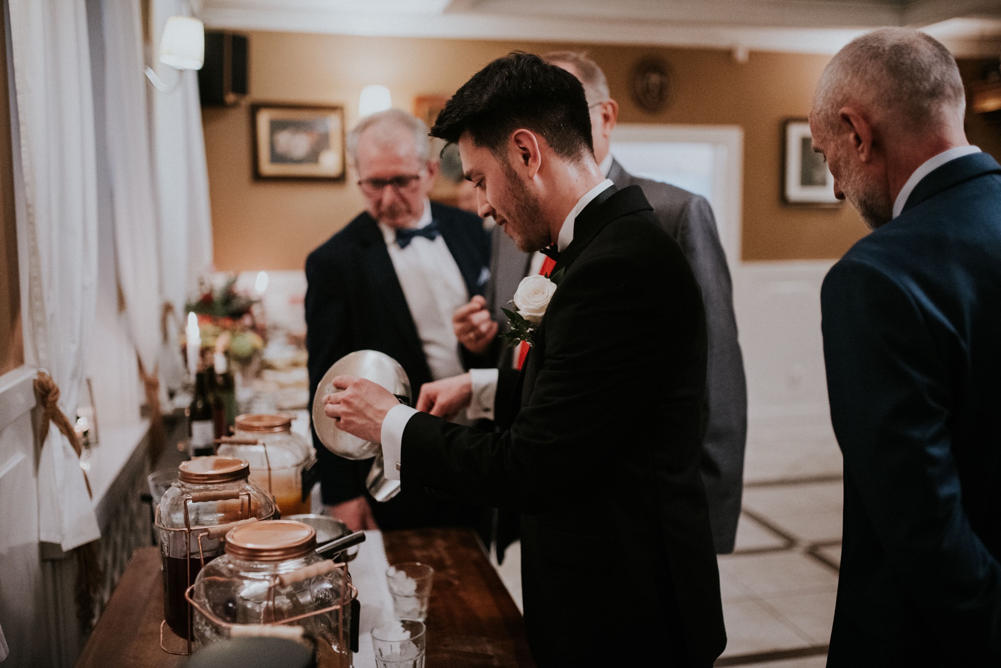 Elegancko ubrany mężczyzna w czarnym smokingu z białą różą w butonierce nalewa napój z metalowego naczynia do szklanki, na drewnianym stole widoczne szklane pojemniki z napojami i inne osoby w tle.