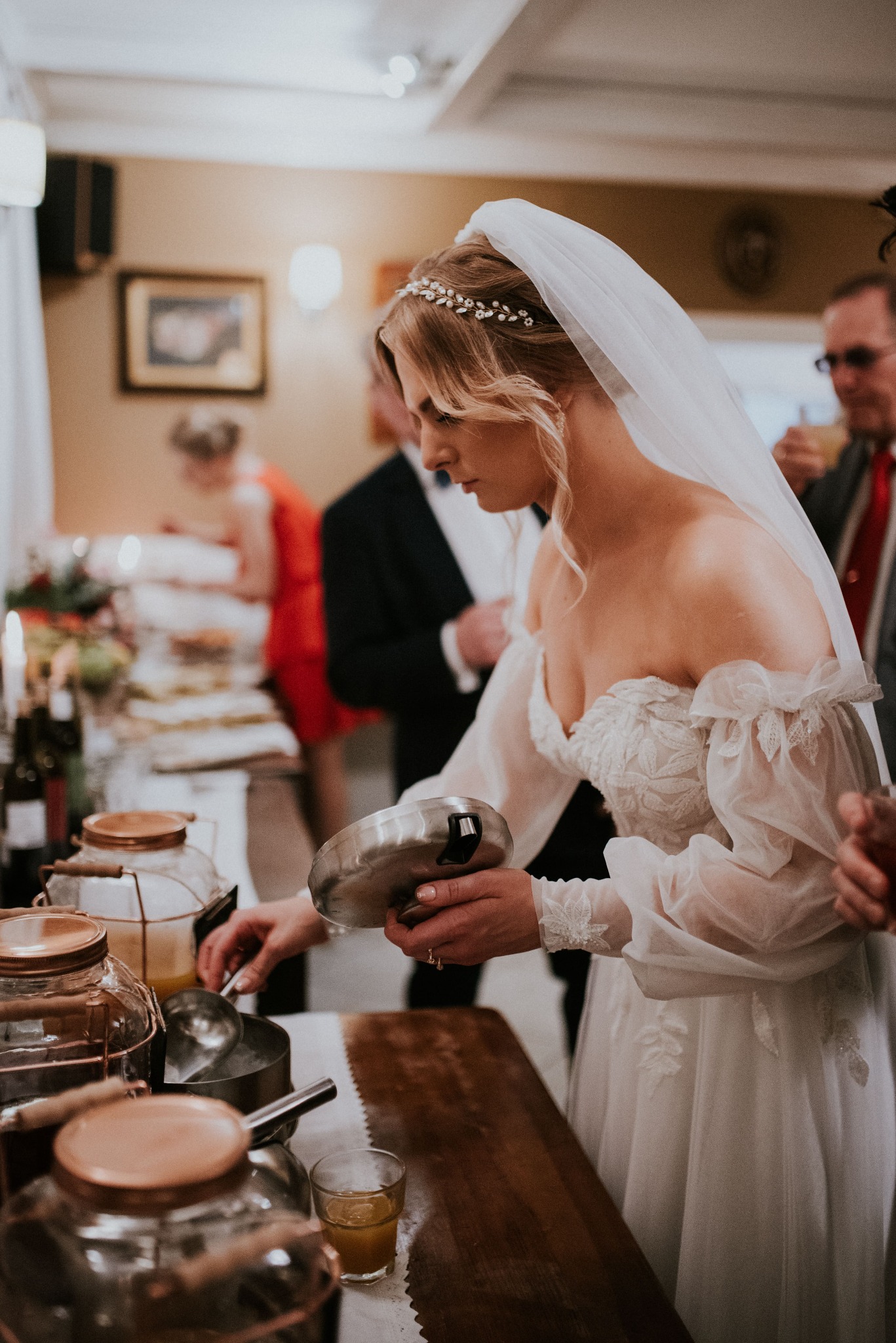 Panna młoda w białej sukni nabiera chochlą napój z metalowej miski na stole z napojami i przekąskami podczas przyjęcia weselnego.