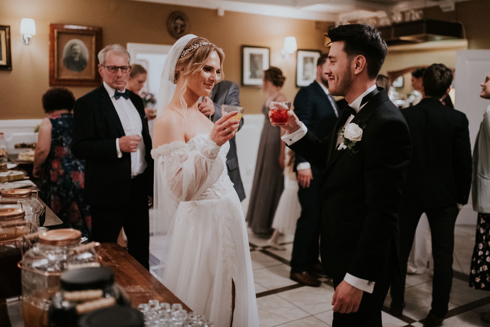 Panna młoda w zwiewnej sukni i pan młody w czarnym smokingu wznoszą toast koktajlami podczas przyjęcia weselnego, w tle goście i bufet z napojami.