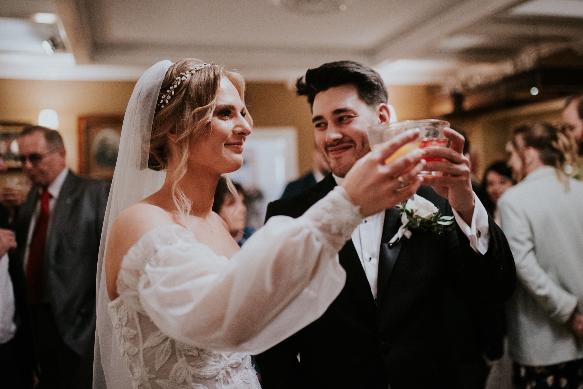 Uśmiechnięta panna młoda w sukni z koronkowymi aplikacjami i welonem oraz pan młody w smokingu wznoszą toast pomarańczowymi koktajlami na tle gości weselnych.