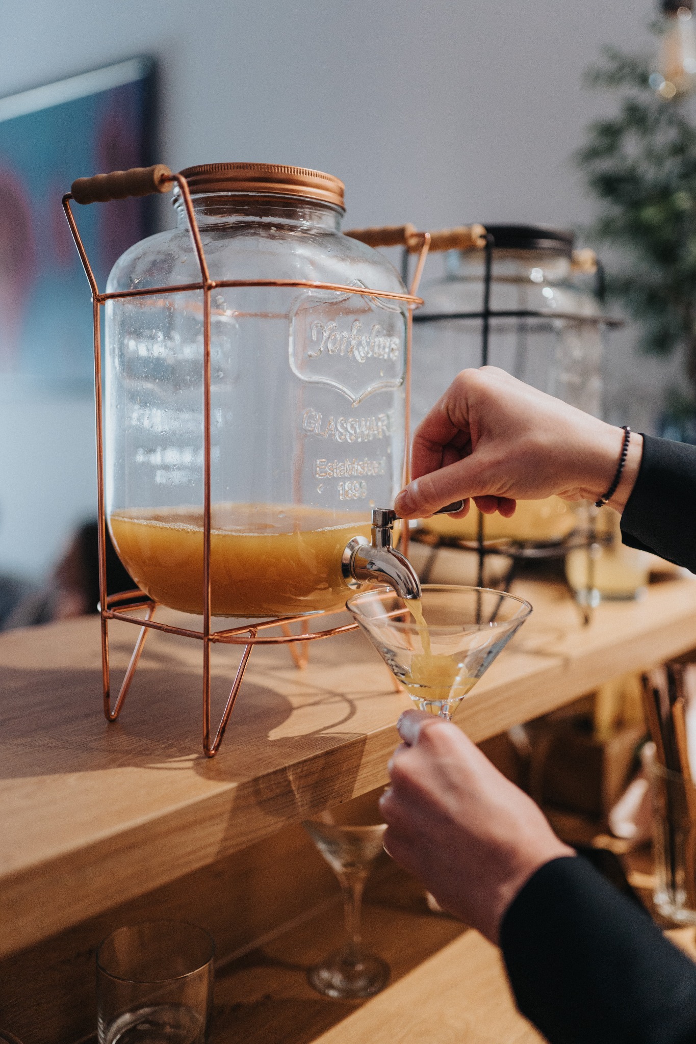 Nalewanie pomarańczowego napoju z szklanego dystrybutora z kranikiem do kieliszka koktajlowego; dystrybutor na metalowym stojaku w kolorze różowego złota.