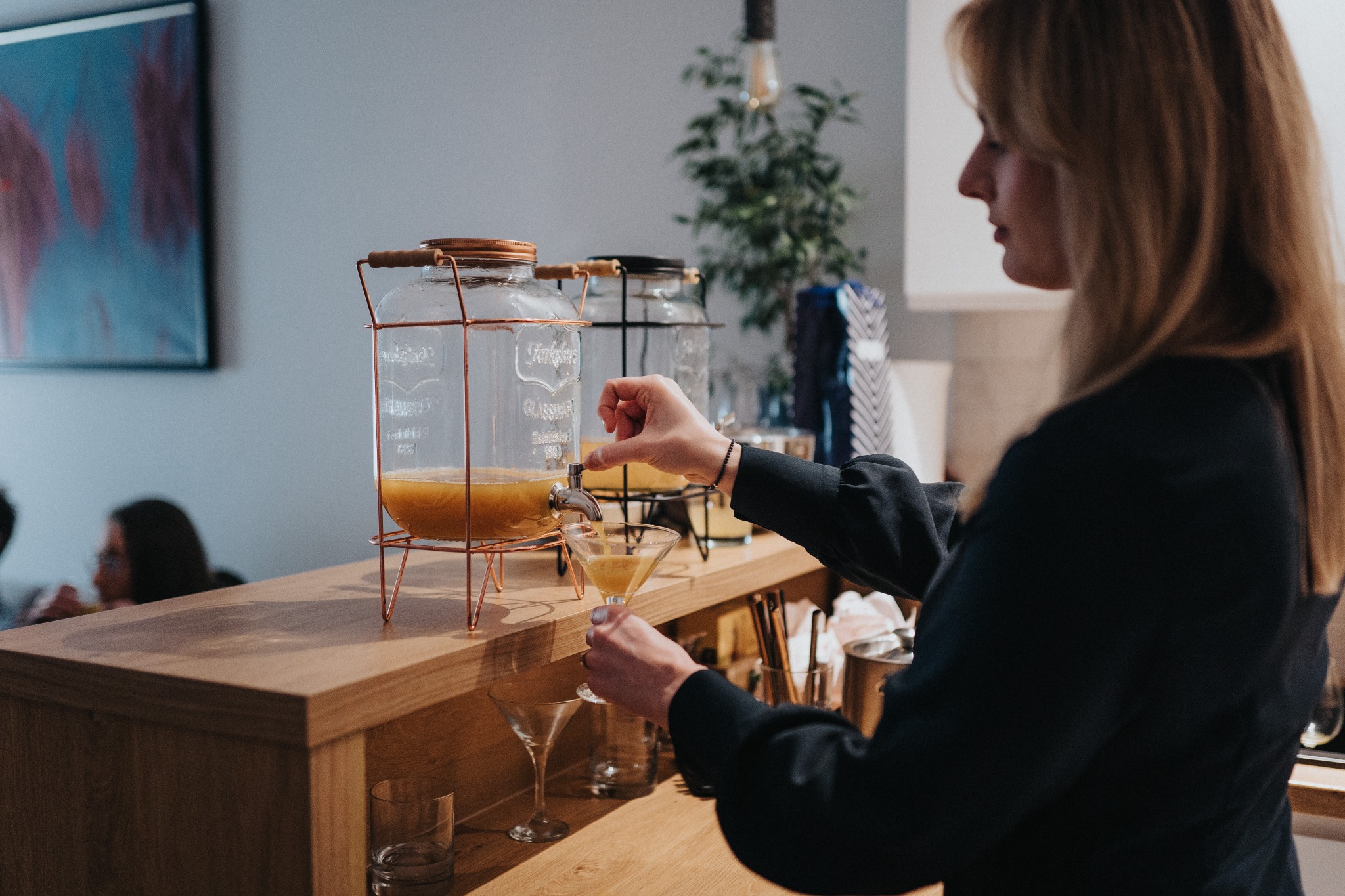 Kobieta nalewa pomarańczowy napój z dużego szklanego dystrybutora z kranikiem do kieliszka koktajlowego na drewnianym barze, widoczne dodatkowe kieliszki i dekoracje.