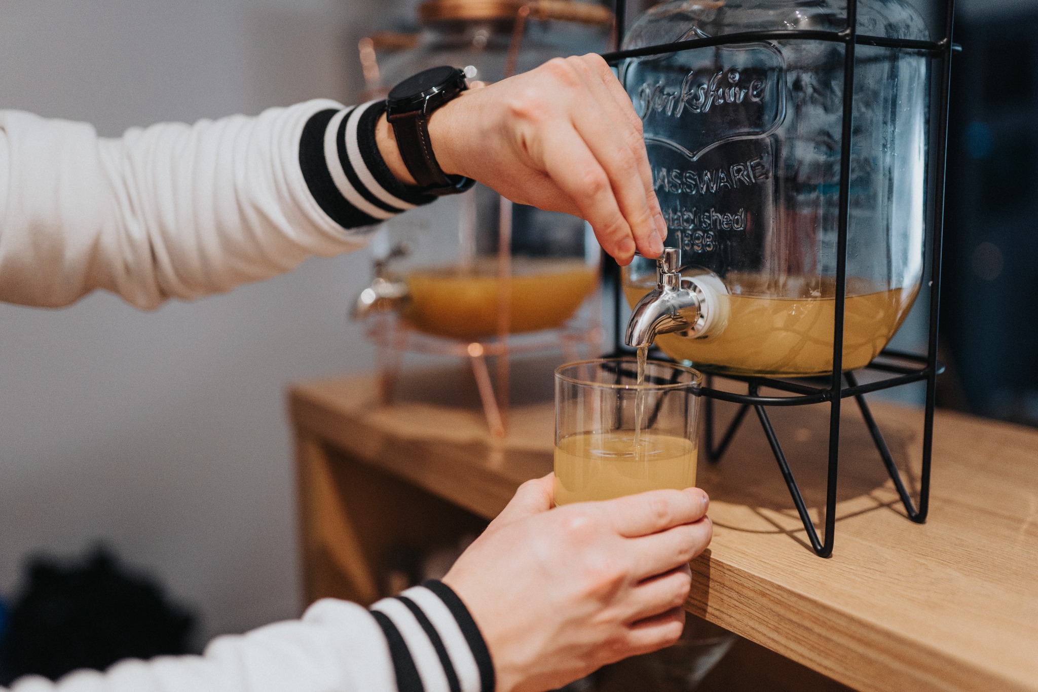 Osoba w bluzie w paski nalewa napój z dużego szklanego dystrybutora z kranikiem do szklanki. W tle widoczny drugi dystrybutor.
