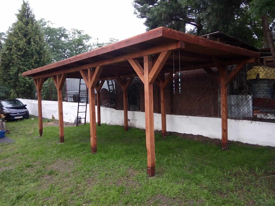 Zadaszona konstrukcja z drewna na trawniku, widoczna drabina oparta o tylną ścianę, zaparkowany samochód w tle.