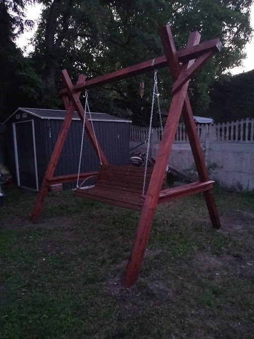 Drewniana huśtawka ogrodowa z siedziskiem na łańcuchach, pomalowana na ciemny kolor, ustawiona na trawniku, w tle widoczny blaszany domek narzędziowy i betonowe ogrodzenie.