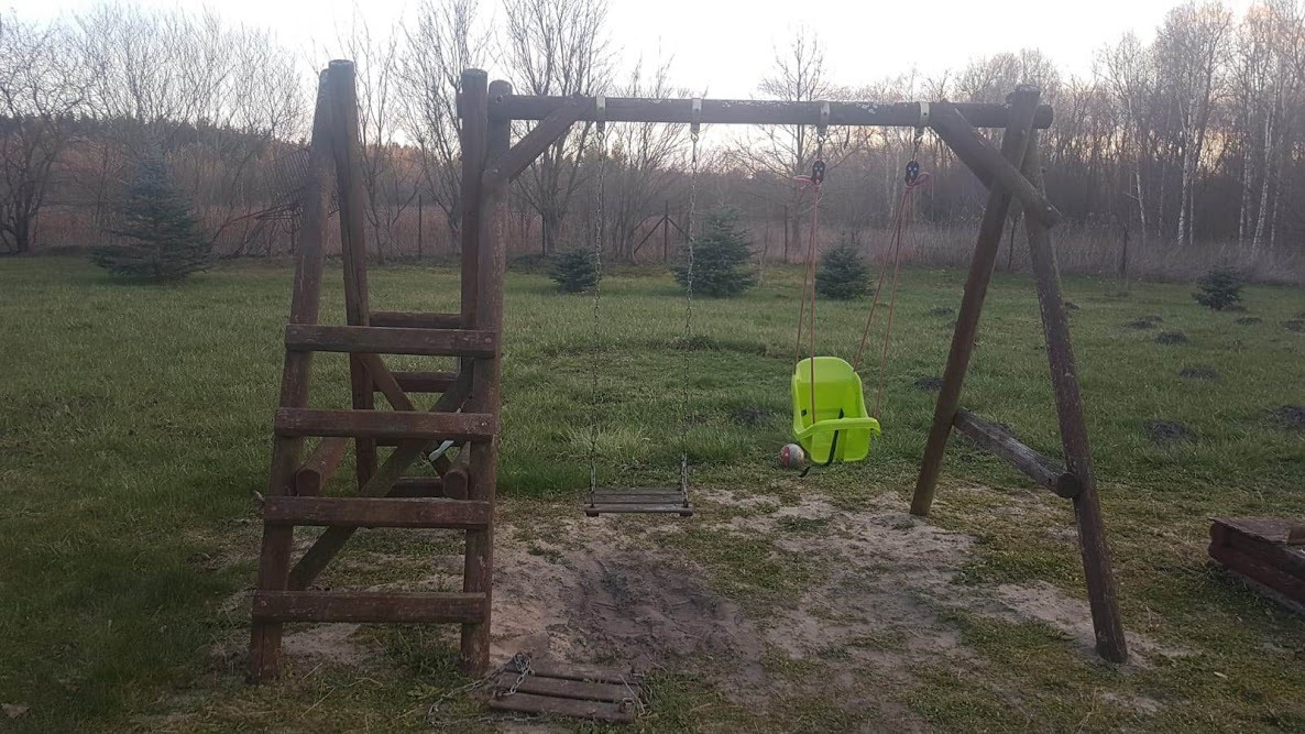Drewniany plac zabaw z huśtawkami na trawiastym podwórku, z widocznymi śladami użytkowania i piłką pod zielonym siedziskiem.