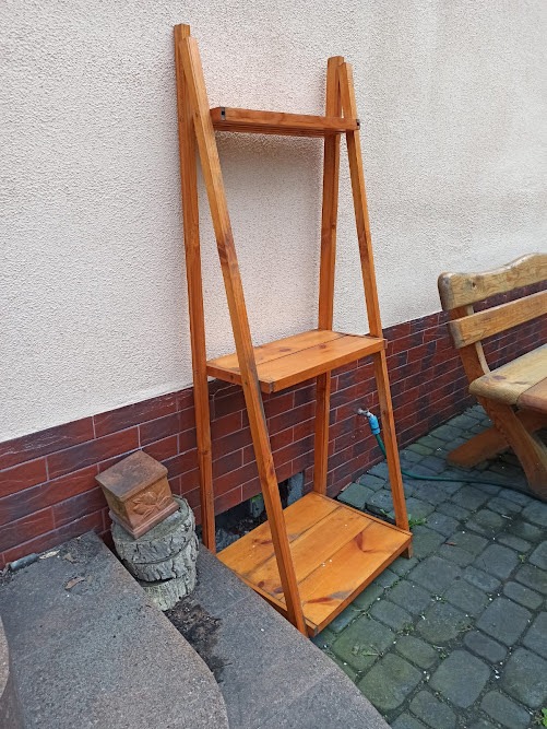 Drewniany regał drabinkowy z trzema półkami, stojący na zewnątrz przy ścianie domu, obok drewniana skrzynka na pniu drzewa i fragment ławki.