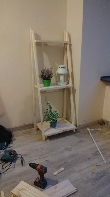 Surowa drewniana półka drabinkowa z roślinami i lampionem, w tle ściana, na podłodze narzędzia: wiertarka, szlifierka, kątownik i elementy drewniane w trakcie montażu.