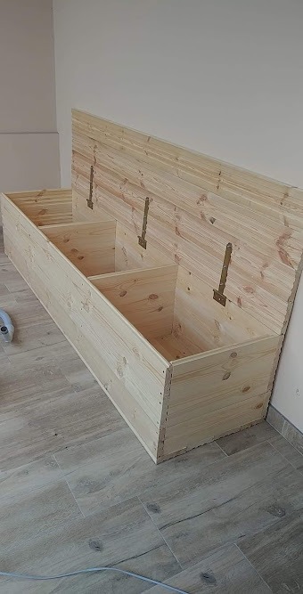 Surowa drewniana skrzynia z trzema przegrodami i podnoszonym wiekiem na tle jasnej ściany i podłogi z płytek imitujących drewno.