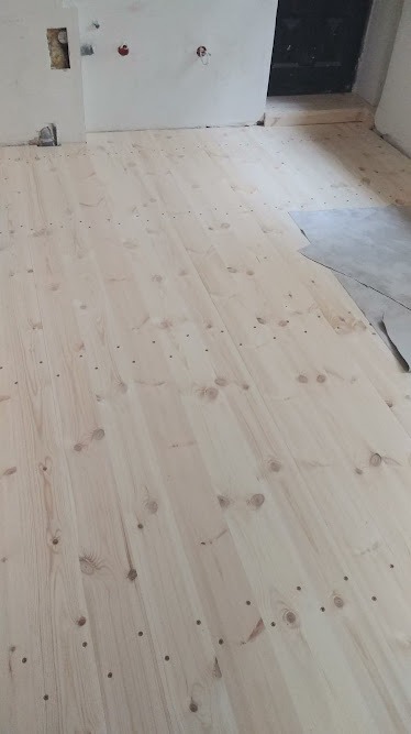 Jasne, surowe deski podłogowe z widocznymi otworami po gwoździach, przygotowane do dalszej obróbki w pomieszczeniu w trakcie remontu.