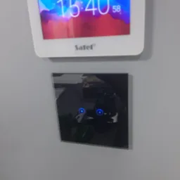 Panel sterowania alarmem Satel z wyświetlaczem LCD i czarnym dotykowym panelem, zamontowany na szarej ścianie.