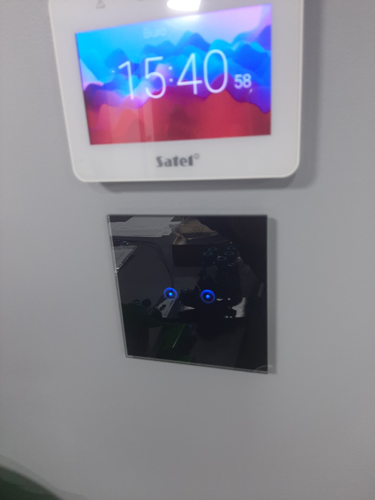 Panel sterowania alarmem Satel z wyświetlaczem LCD i czarnym dotykowym panelem, zamontowany na szarej ścianie.