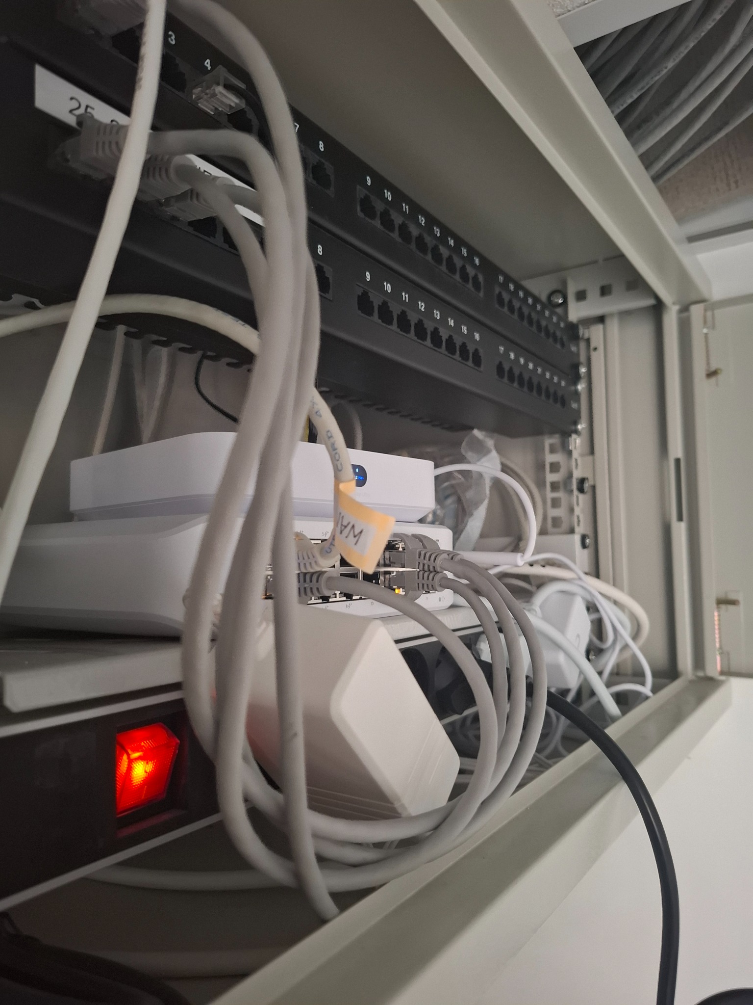 Wnętrze szafy telekomunikacyjnej z patch panelem, routerami i przewodami Ethernet, widoczny czerwony włącznik zasilania. Przewody oznaczone etykietami.