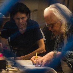 auernes - Dwie osoby pochylone nad dokumentami, jedna pisząca ołówkiem, druga obserwująca, na stole narzędzia i materiały, w tle nieostra rzeźba.