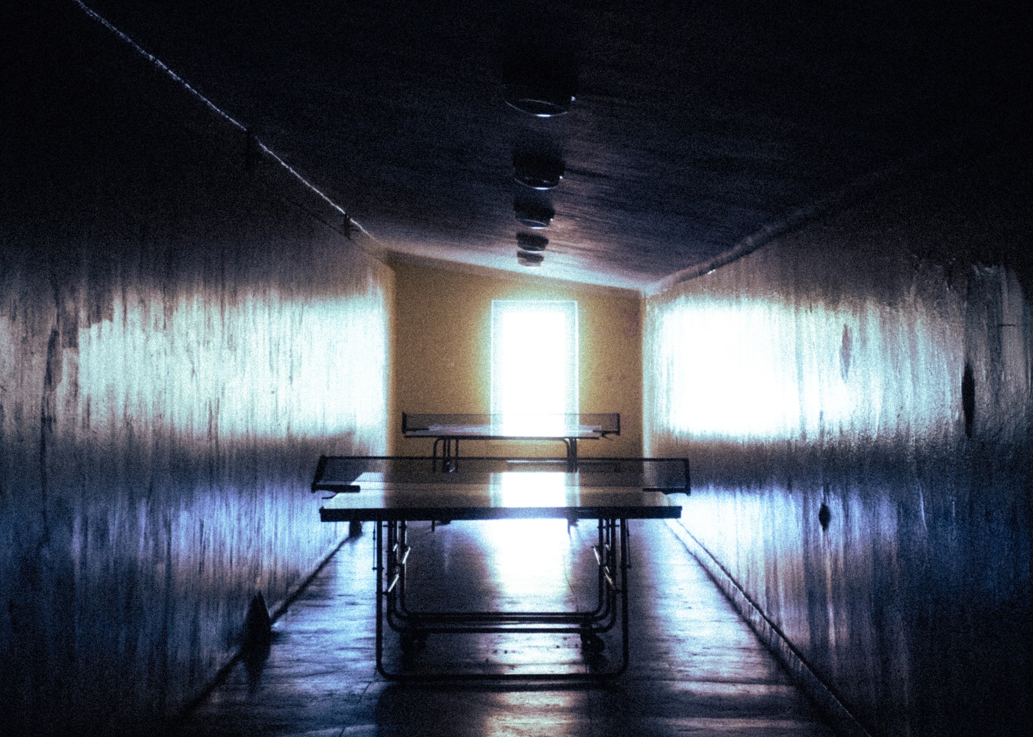 Długi korytarz z betonowymi ścianami prowadzący do jasnego okna, na pierwszym planie stół do ping-ponga ustawiony wzdłuż osi korytarza.