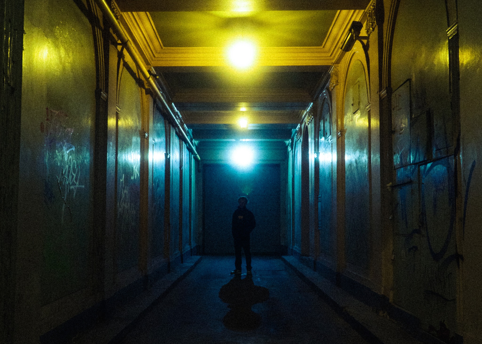 Postać w ciemnym tunelu z graffiti na ścianach, oświetlonym przez górne lampy tworzące atmosferyczny nastrój.