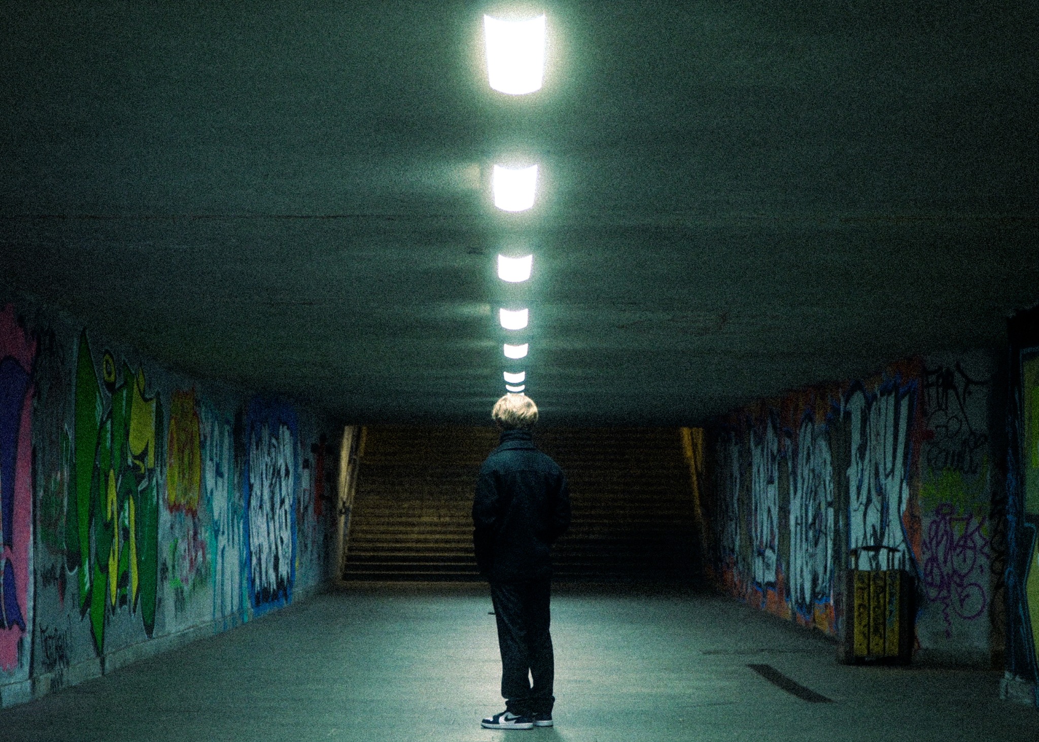 Człowiek stojący w tunelu z graffiti, oświetlonym rzędem lamp sufitowych, ubrany w ciemny płaszcz i sportowe buty.