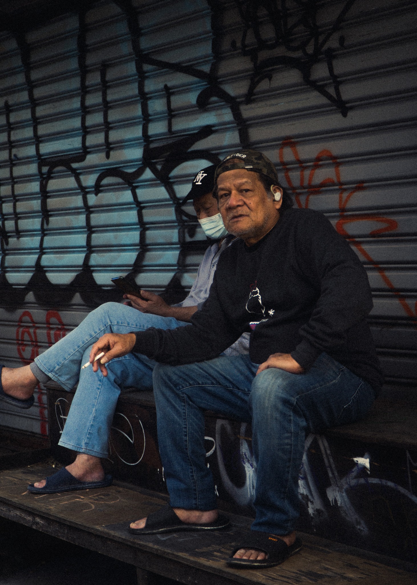 Mężczyzna w czarnej bluzie i dżinsach siedzi na ławce z papierosem w dłoni, obok siedzi osoba w maseczce korzystająca z telefonu, na tle metalowej ściany pokrytej graffiti.