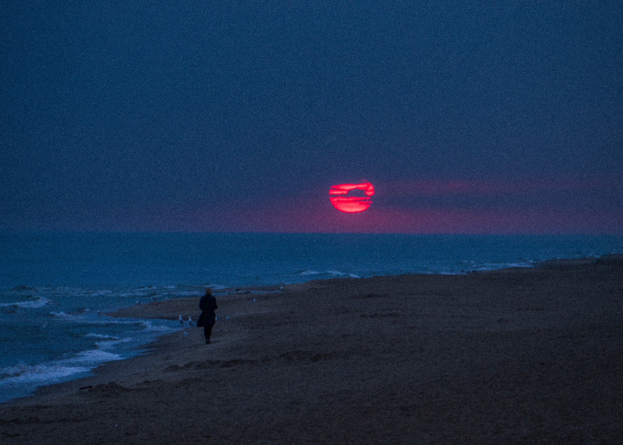 Sylwetka kobiety w płaszczu spacerującej po plaży o zmierzchu, z czerwonym słońcem zachodzącym nad morzem i falami rozbijającymi się o brzeg.