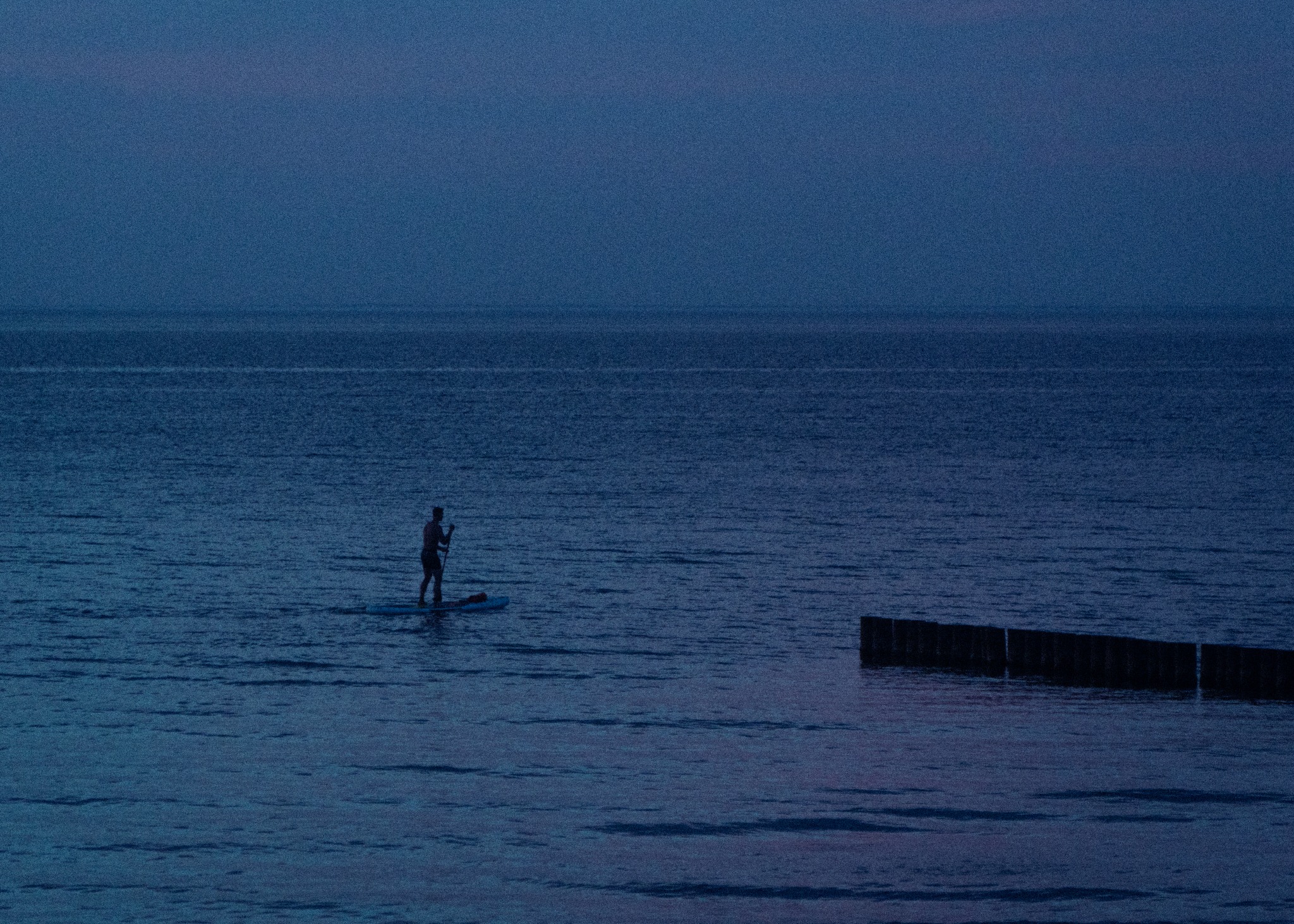 Sylwetka osoby pływającej na desce SUP na spokojnym morzu o zmierzchu, widoczny falochron w oddali.