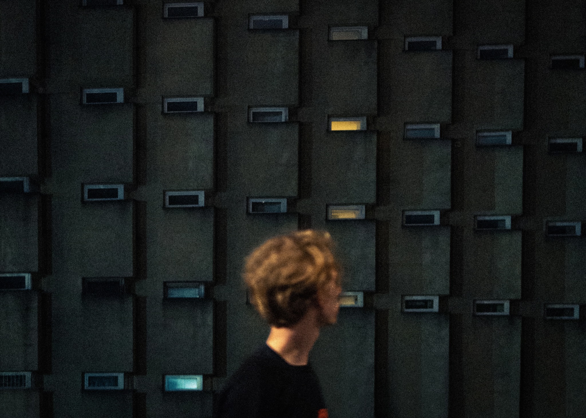 Mężczyzna z rozmyciem ruchu na tle brutalistycznej fasady budynku z małymi oknami, z których niektóre są oświetlone od wewnątrz.