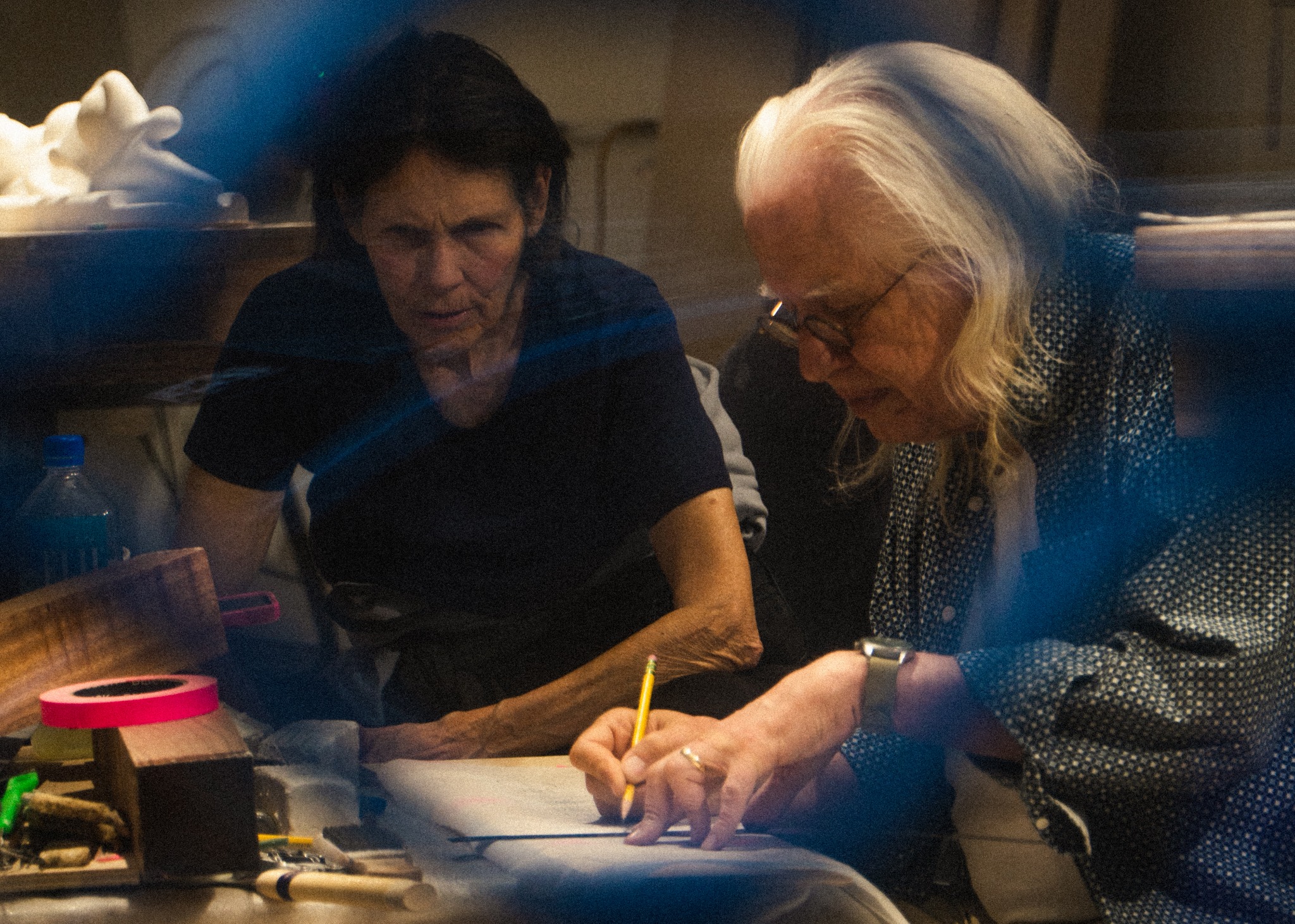 Dwie osoby pochylone nad dokumentami, jedna pisząca ołówkiem, druga obserwująca, na stole narzędzia i materiały, w tle nieostra rzeźba.