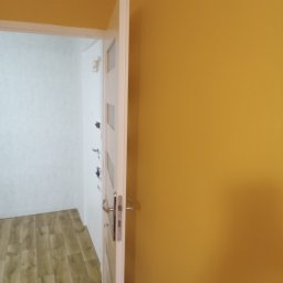 System Perfect Sp. z o.o. - Otwarta biała ościeżnica drzwiowa z widoczną ścianą pomalowaną na musztardowy kolor i fragmentem podłogi z paneli drewnopodobnych w tle.