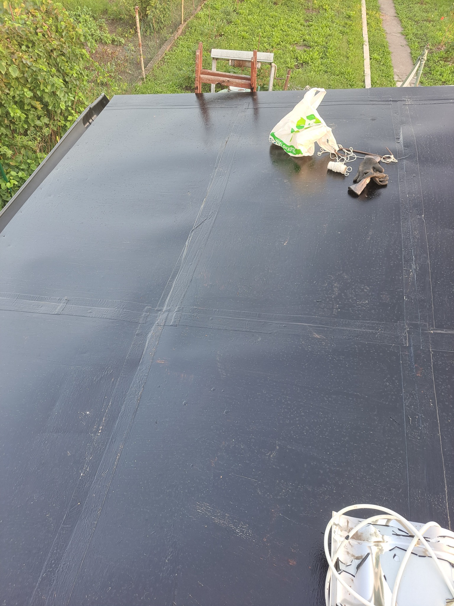 Czarna, płaska powierzchnia dachu pokryta papą, z widocznymi łączeniami i narzędziami pracy, torba foliowa i lampa na wierzchu, w tle zieleń i elementy konstrukcyjne.