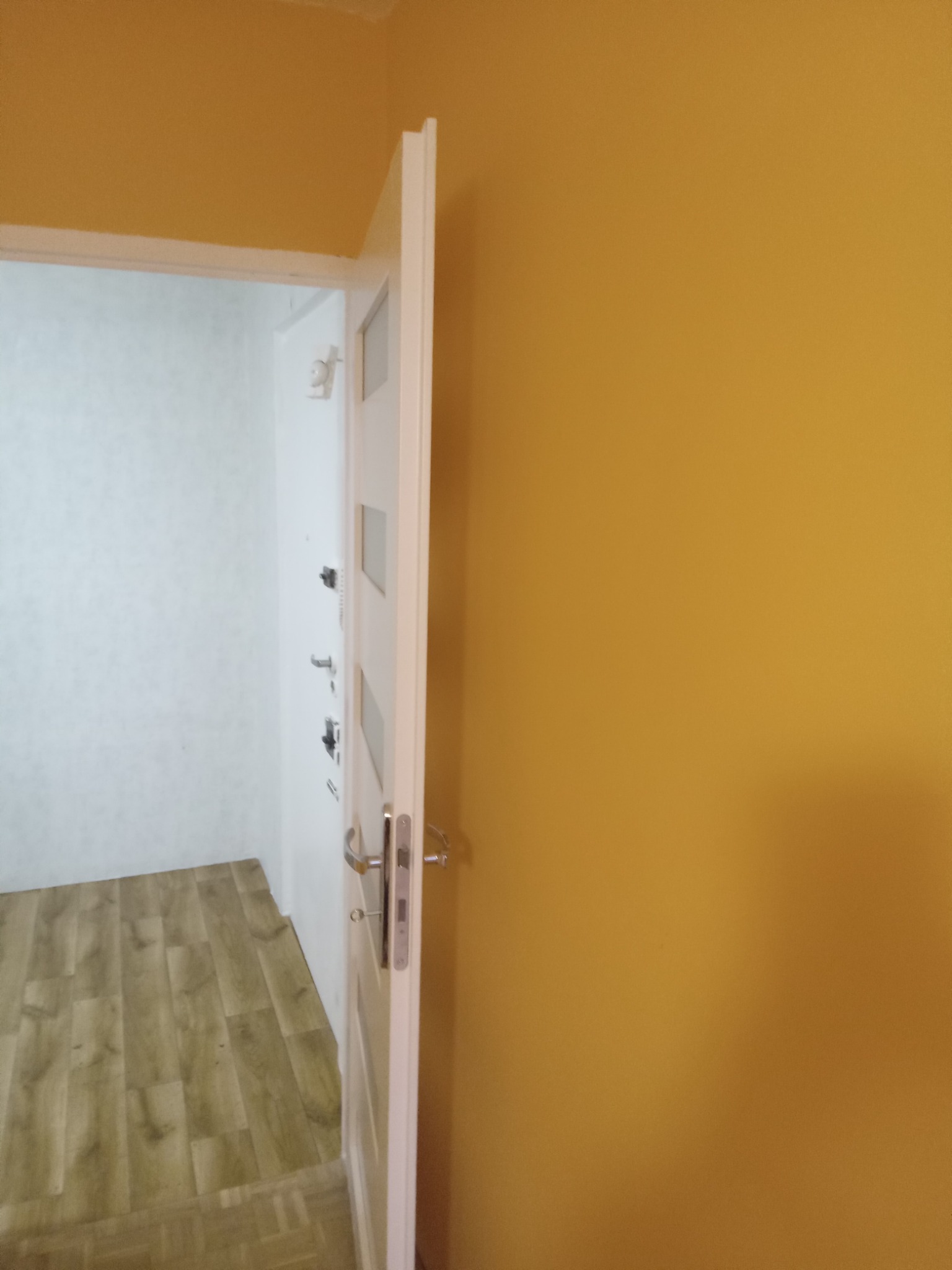 Otwarta biała ościeżnica drzwiowa z widoczną ścianą pomalowaną na musztardowy kolor i fragmentem podłogi z paneli drewnopodobnych w tle.