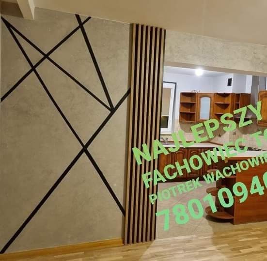 Ściana z szarą fakturą i geometrycznym wzorem z czarnych linii, obok pionowy panel z drewnianych listewek. W tle widoczny fragment kuchni z drewnianymi szafkami. Na zdjęciu widoczne napisy reklamowe.