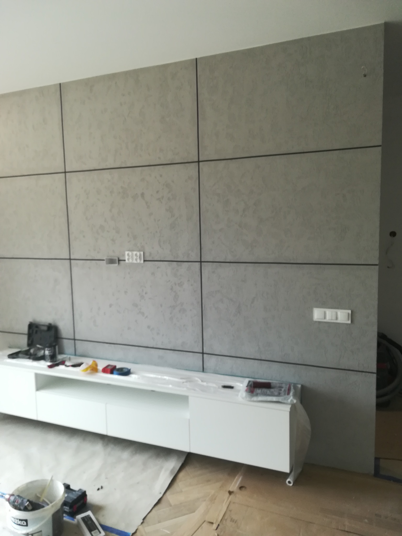 Ściana z panelami dekoracyjnymi w trakcie montażu, widoczne narzędzia i zabezpieczenia podłogi, surowe wykończenie.