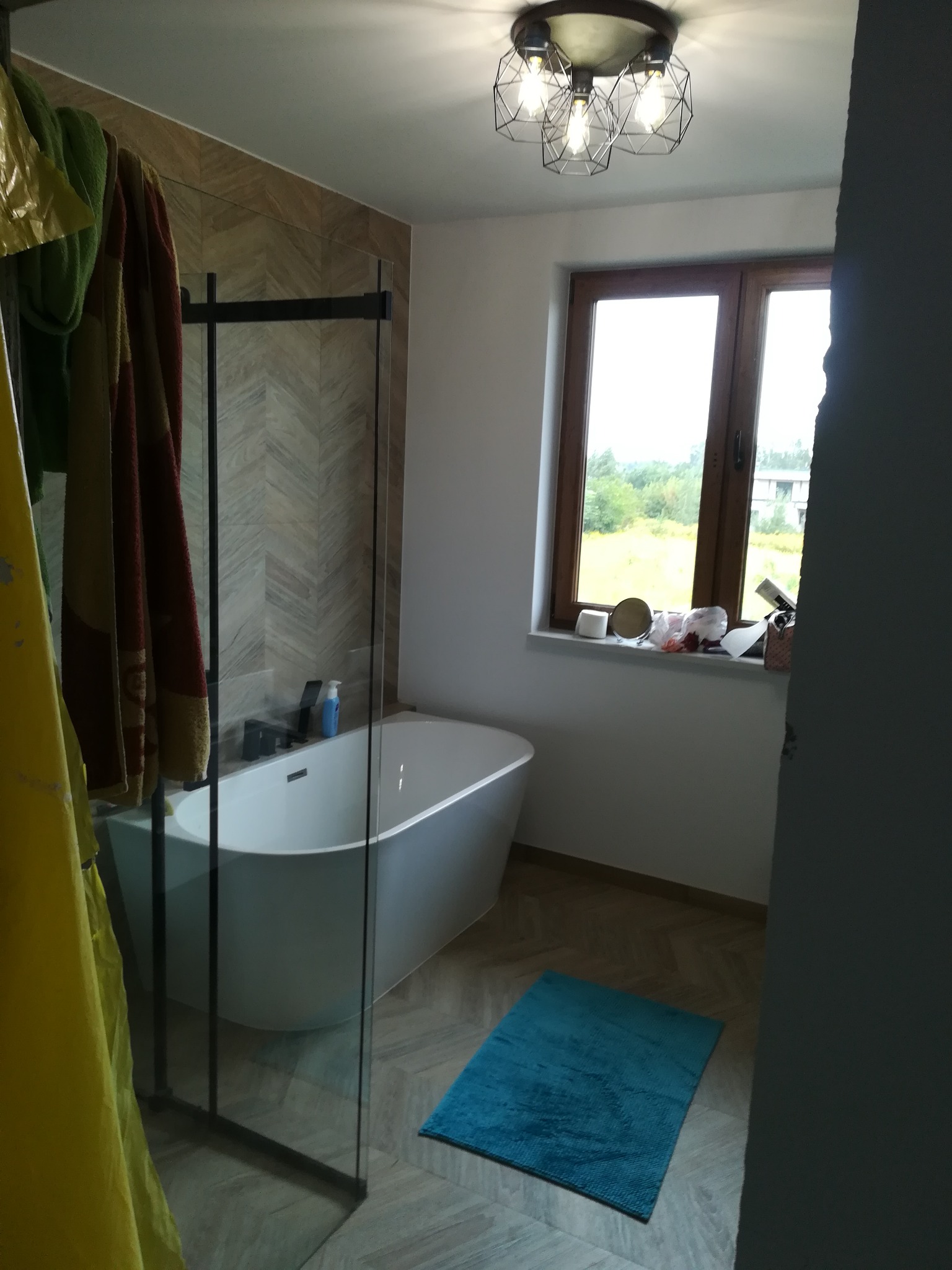 Nowoczesna łazienka z wolnostojącą białą wanną za szklaną ścianką prysznicową, wykończona płytkami w jodełkę, z oknem i dekoracyjną lampą sufitową, oraz niebieskim dywanikiem.