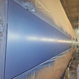 BTT Polska - Duży element konstrukcyjny, prawdopodobnie pojazdu, po nałożeniu świeżej, gładkiej, niebieskiej powłoki lakierniczej w kabinie lakierniczej, z widocznymi śladami zabezpieczeń i taśmy malarskiej.