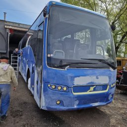 BTT Polska - Niebieski autobus w trakcie renowacji lakierniczej, z widocznymi śladami szpachli i taśmy maskującej, na tle warsztatu i drzew.