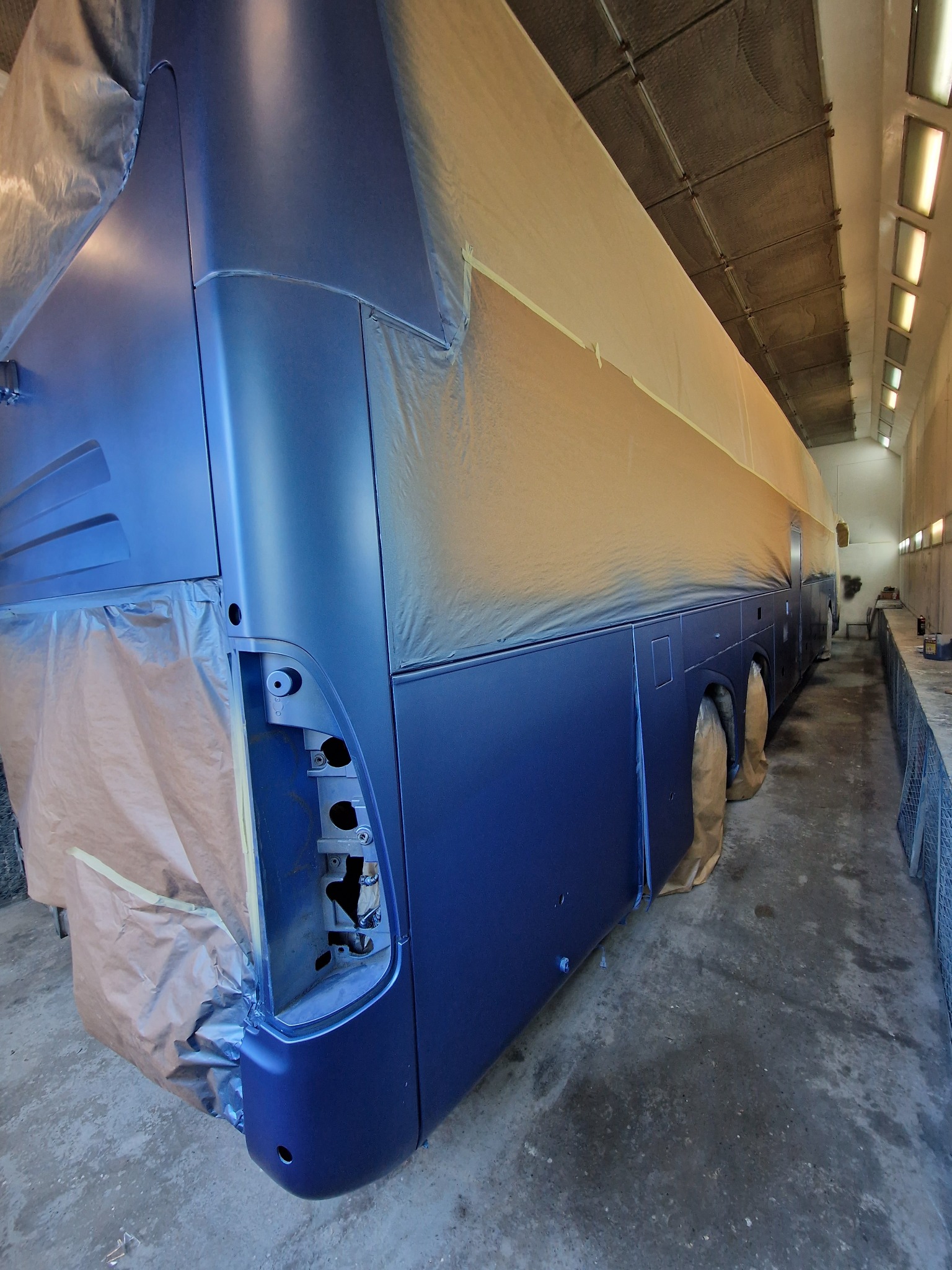 Autobus w trakcie lakierowania w kabinie lakierniczej, zabezpieczony folią malarską, świeżo pomalowany na ciemnoniebieski kolor.