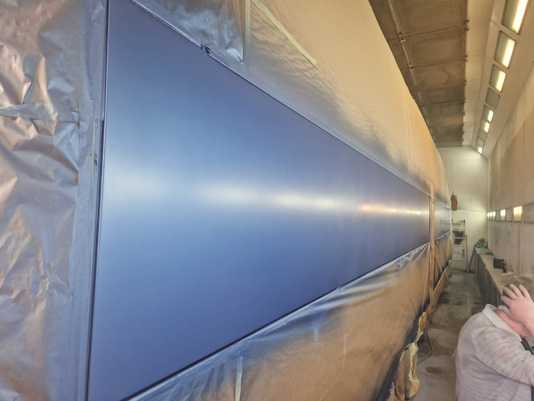 Duży element konstrukcyjny, prawdopodobnie pojazdu, po nałożeniu świeżej, gładkiej, niebieskiej powłoki lakierniczej w kabinie lakierniczej, z widocznymi śladami zabezpieczeń i taśmy malarskiej.