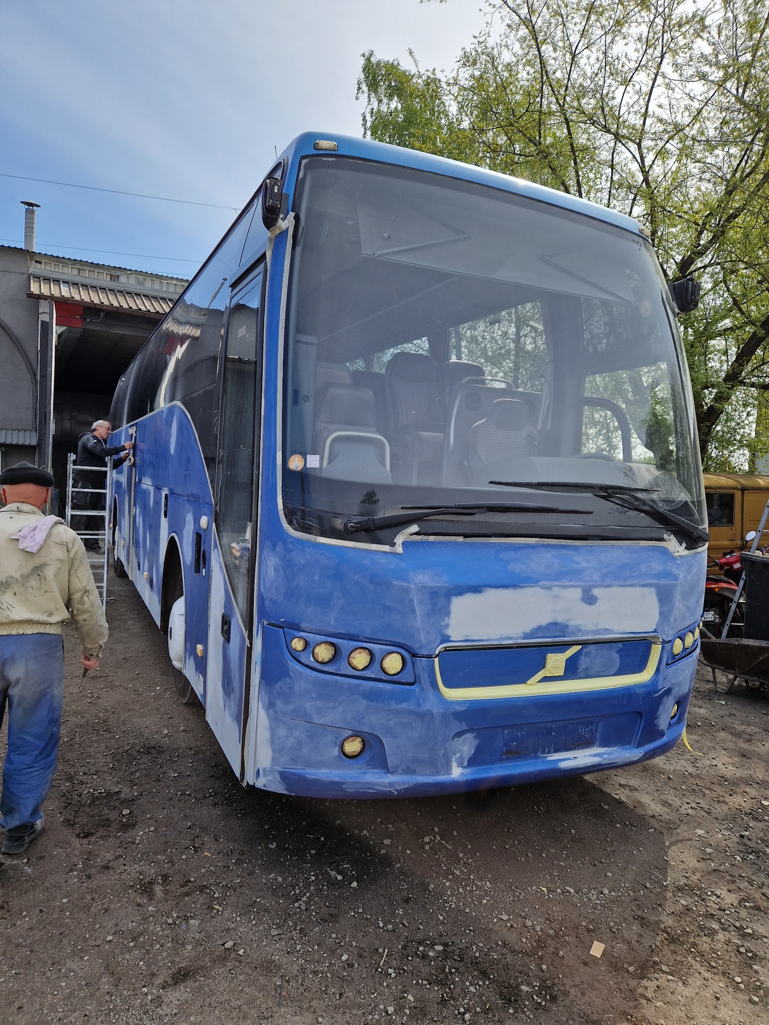 Niebieski autobus w trakcie renowacji lakierniczej, z widocznymi śladami szpachli i taśmy maskującej, na tle warsztatu i drzew.