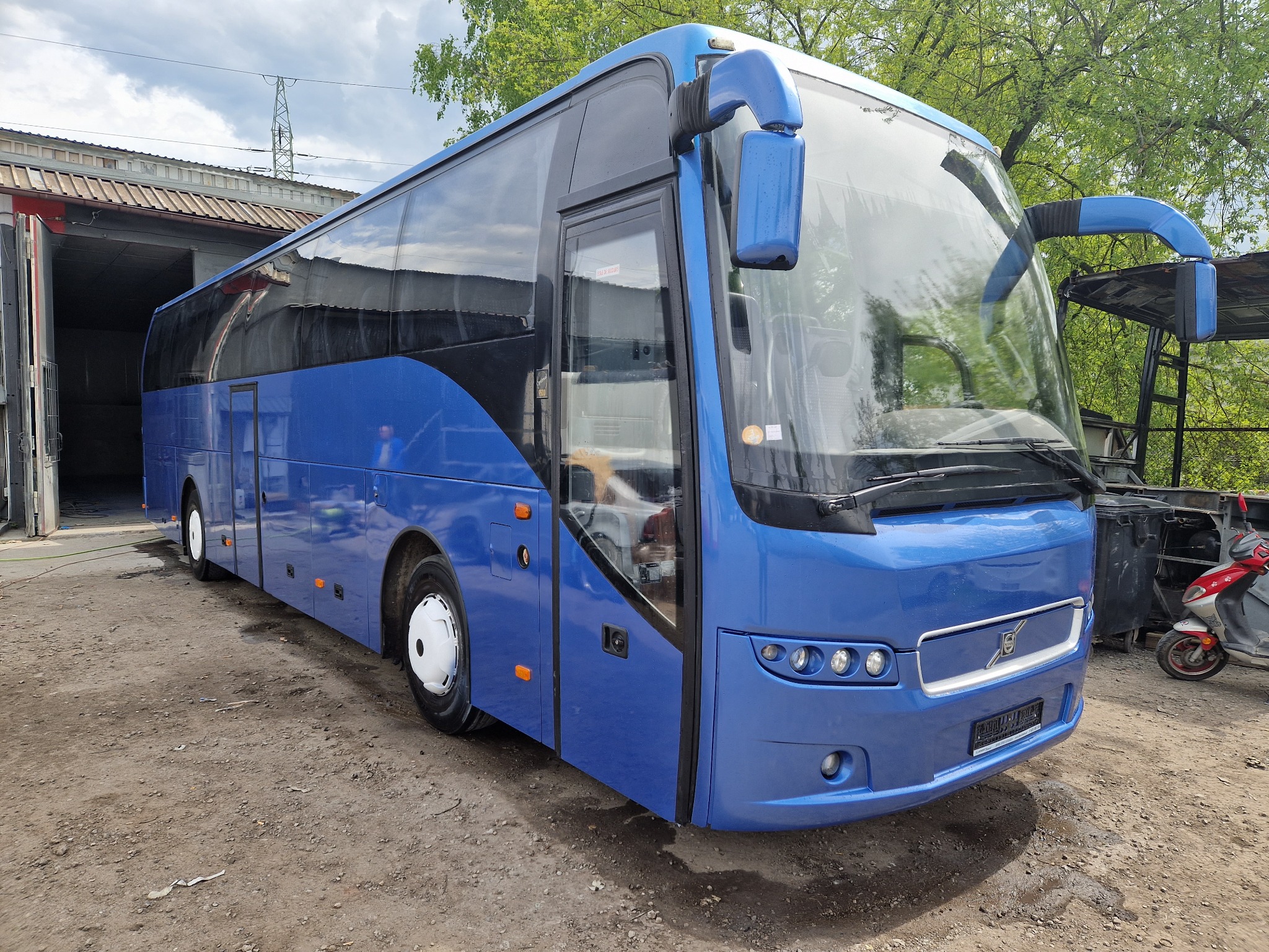 Niebieski autobus marki Volvo po odnowieniu lakieru, stojący na placu przed budynkiem warsztatowym z otwartymi wrotami, widoczny skuter w tle.