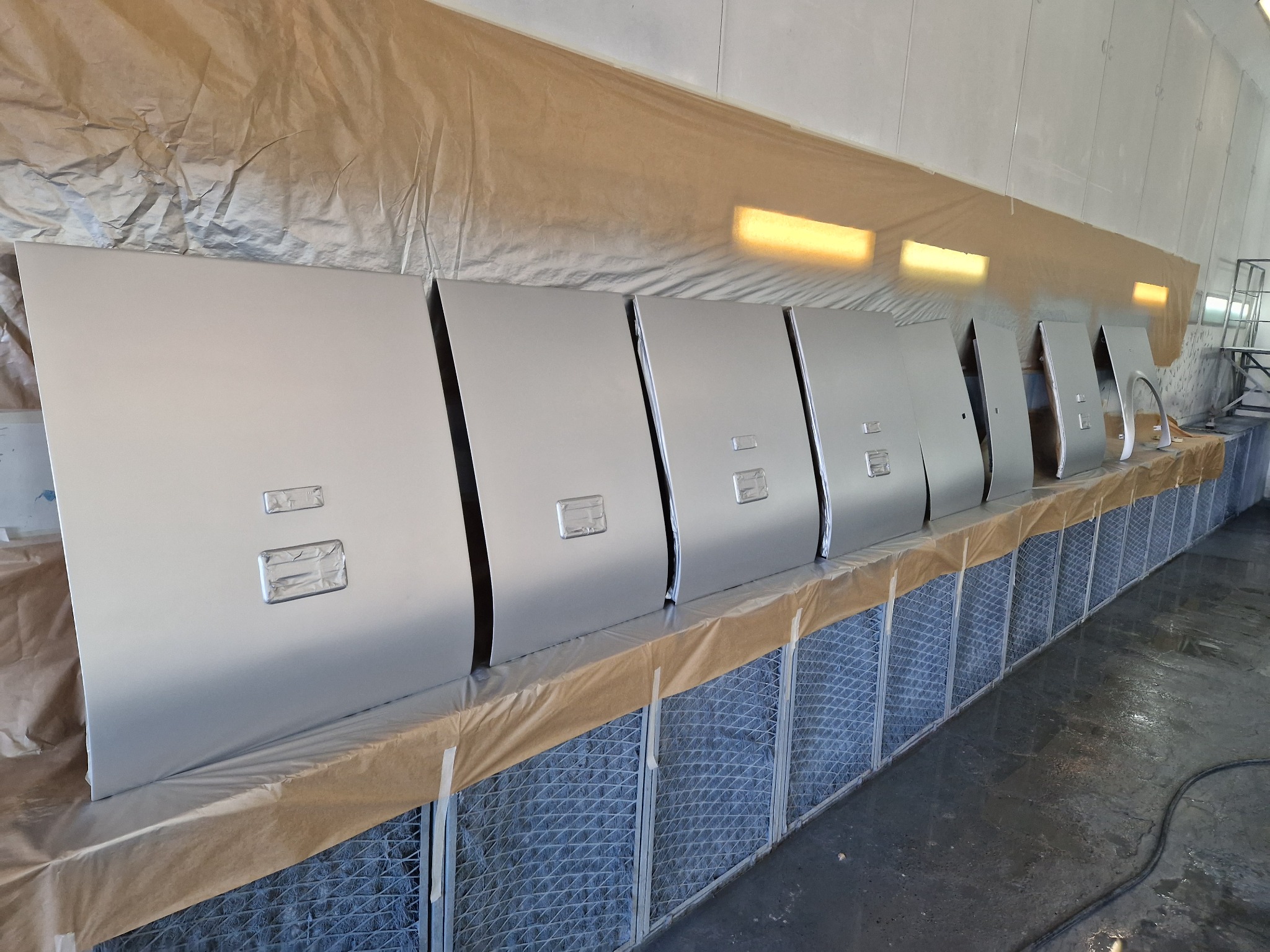 Pomalowane na srebrno drzwi samochodowe i błotniki ustawione w rzędzie w kabinie lakierniczej, zabezpieczone papierem i folią, widoczne filtry powietrza w dolnej części kabiny.