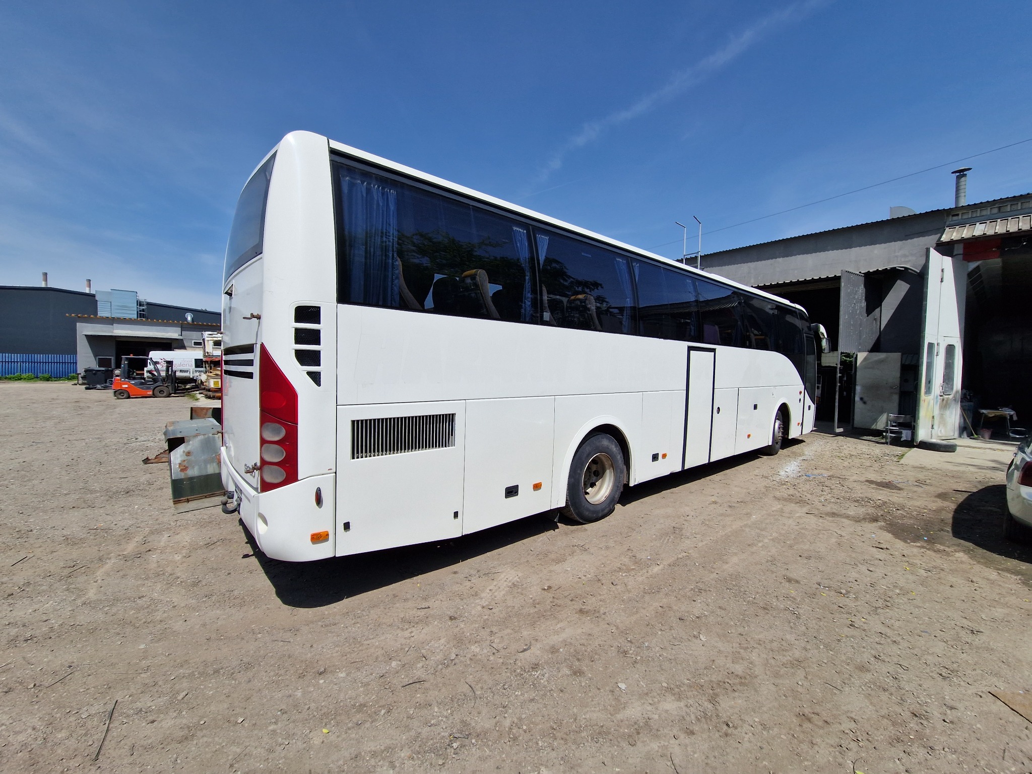 Biały autokar po renowacji lakierniczej, stojący na placu warsztatowym z widocznymi śladami użytkowania w tle, w słoneczny dzień.