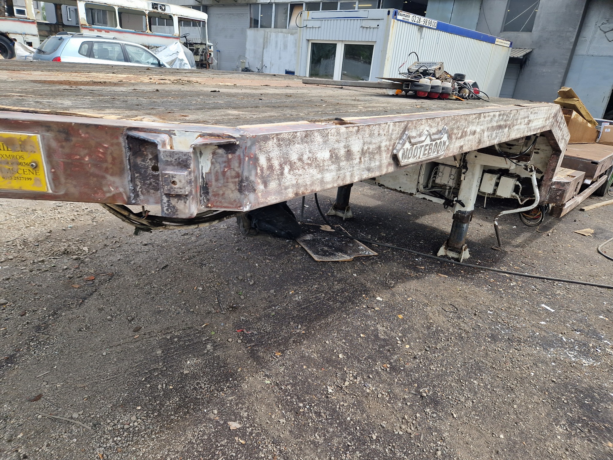 Przyczepa samochodowa z widocznymi śladami korozji i wcześniejszej obróbki, z żółtą tabliczką informacyjną i fragmentami starej farby, stojąca na szarym betonowym podłożu.