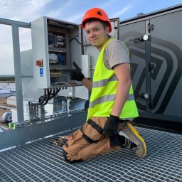 OSW Pro - Uśmiechnięty elektryk w kasku i kamizelce, klęczy obok otwartej szafy sterowniczej na dachu, pokazując kciuk w górę. Widoczne narzędzia i instalacja elektryczna.