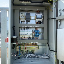 OSW Pro - Otwarta, szara szafa sterownicza z widocznymi elementami elektrycznymi: gniazdo, bezpieczniki, przekaźniki, listwy zaciskowe i przewody. Widok z bliska na instalację.