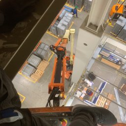 OSW Pro - Widok z podnośnika koszowego na halę produkcyjną podczas prac na wysokościach. Widoczny pracownik w oddali i elementy konstrukcyjne hali.