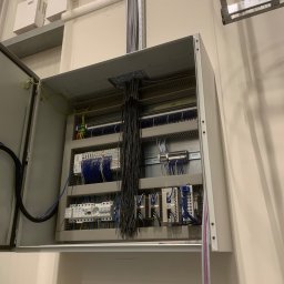 OSW Pro - Otwarta szafa sterownicza z widocznymi elementami instalacji elektrycznej, przewodami i zabezpieczeniami. Widoczna kamera przemysłowa i kable w peszlu.